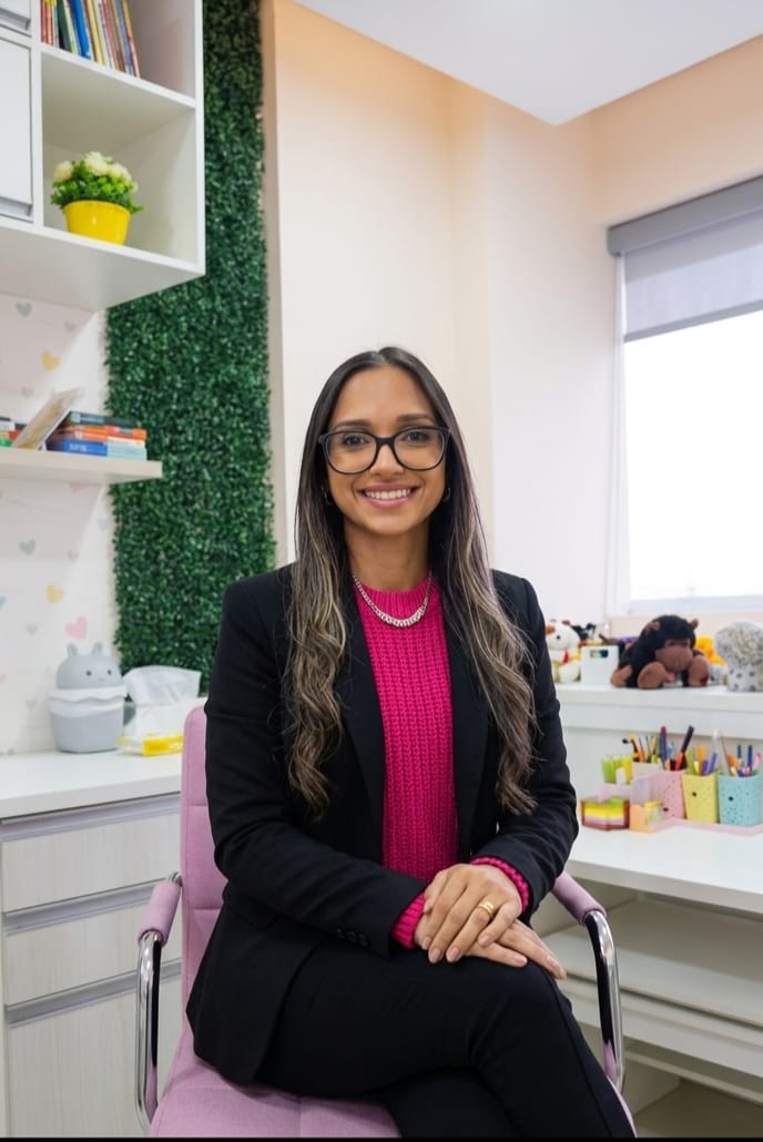 Aline Andolfo sentada no consultório, com a parede de folhagem e os brinquedos ao fundo